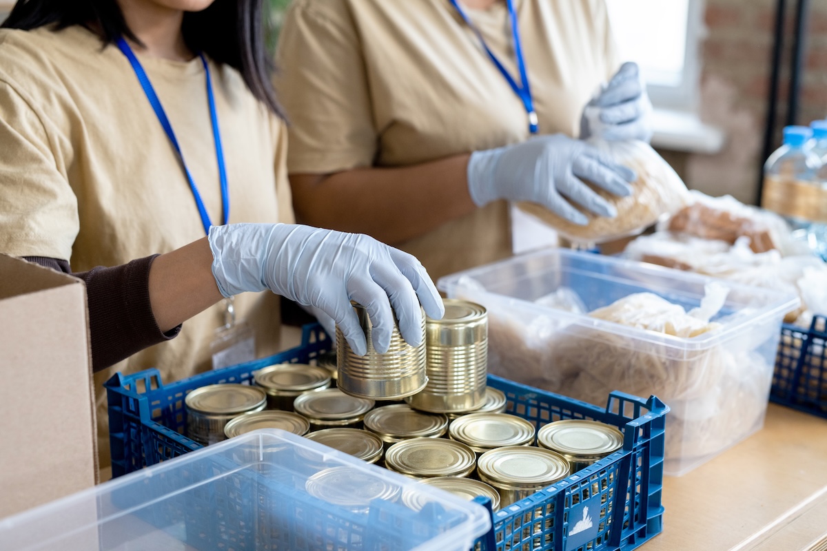 Gloved hand of female volunteer putting tin can containing steamed meat from plastic box